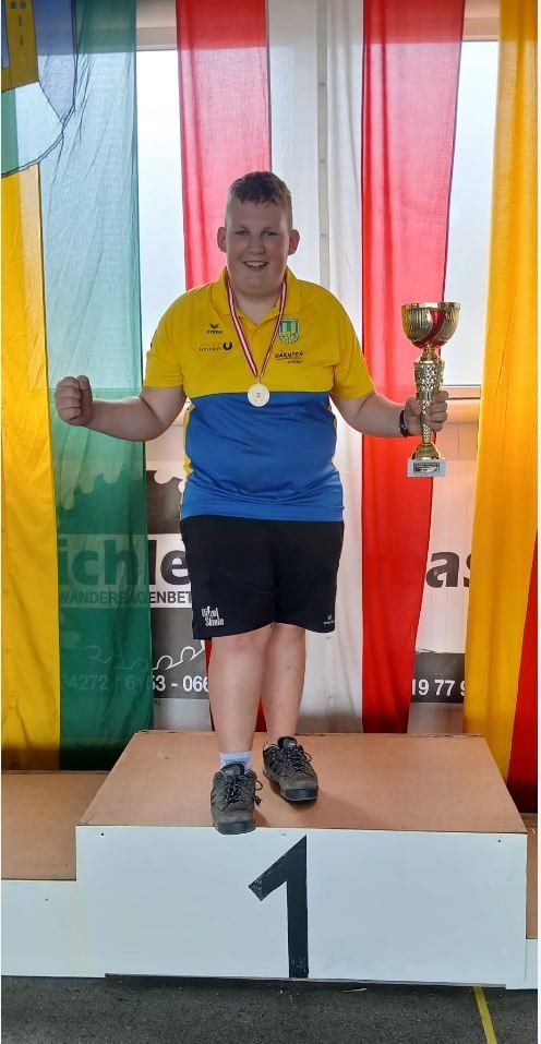 Ein junger Mann mit einer Medaille um den Hals steht mit einem Pokal auf einem Podest, lächelt und hat Flaggen im Hintergrund.