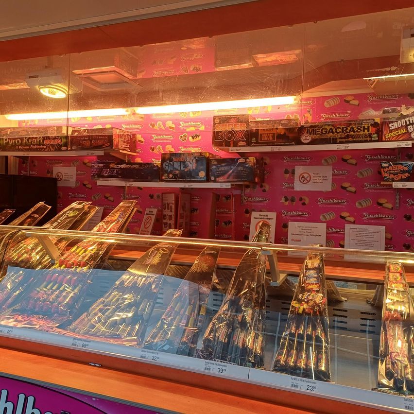 Ein Geschäft zeigt eine Vielzahl von Feuerwerkskörpern in einer Glasvitrine. Rosa Wände mit verschiedenen Marken und Produkten. Viele bunte Kisten mit Feuerwerkskörpern und Süßigkeiten schmücken die Regale. Ein helles Licht beleuchtet die Produkte.