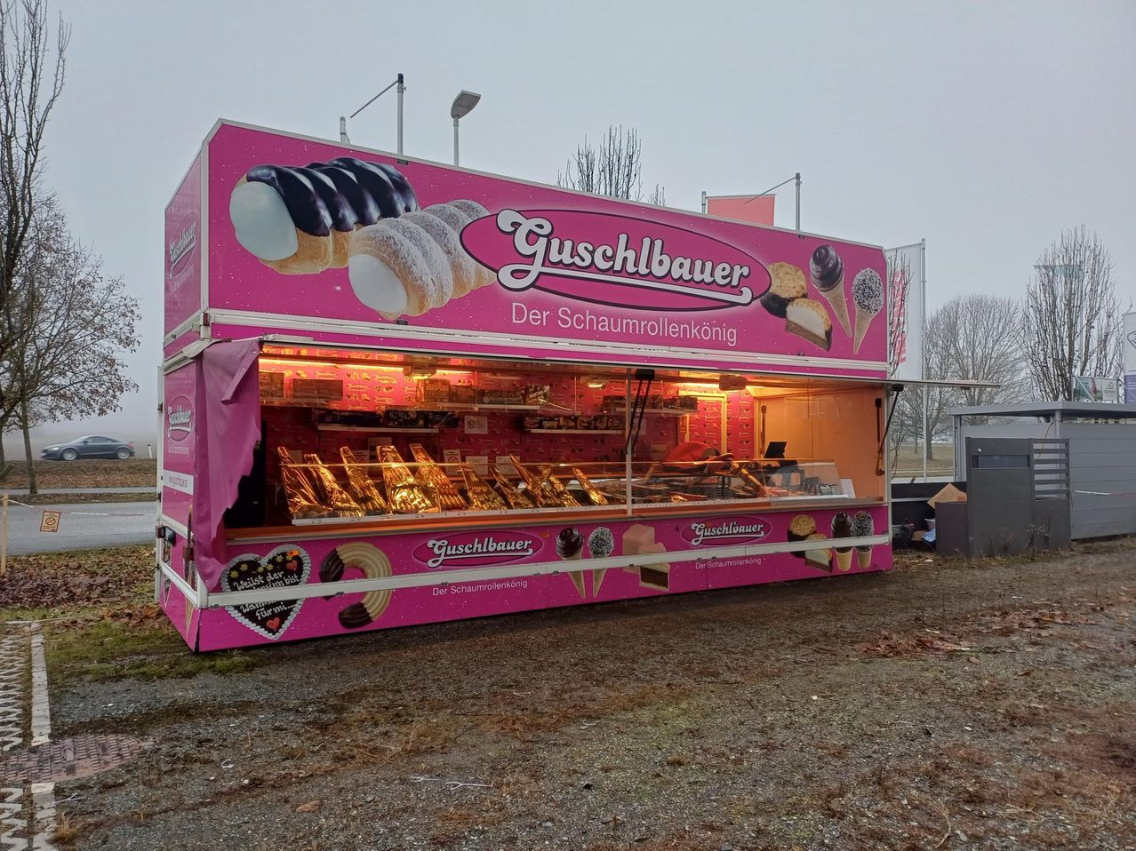 Ein rosafarbener Imbisswagen mit dem Namen Guschbauer an der Seite, der verschiedene Desserts anbietet, darunter Croissants und Eisbecher.