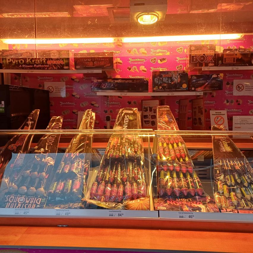 Eine Ladenvitrine zeigt verschiedene Feuerwerke und Süßigkeiten. Im Vordergrund befinden sich mehrere Packungen von Feuerwerken mit Etiketten, die verschiedene Größen angeben. Hinter der Vitrine sind Regale mit weiteren Feuerwerken und Süßigkeiten gefüllt. Der Laden hat eine pinke Wand, die mit Herzdesigns und Produktetiketten verziert ist.