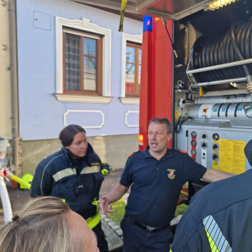 Zwei Feuerwehrleute und ein Mann in einem blauen Hemd stehen vor einem Feuerwehrauto mit einer Leiter.