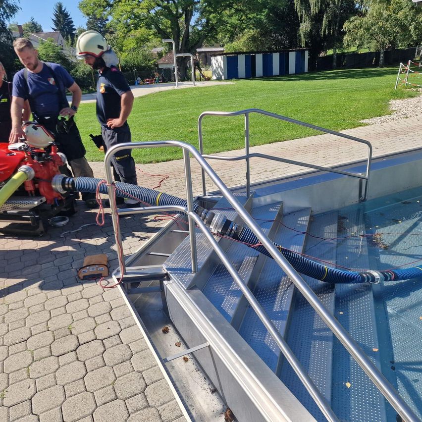 Zwei Feuerwehrleute stehen neben einer Pumpe, die mit einem Schlauch verbunden ist, in der Nähe eines Pools. Ein Mann schaut sich die Pumpe an. Der Pool hat eine Metallhandlauf. Im Hintergrund befindet sich eine Rasenfläche mit einem Gebäude.