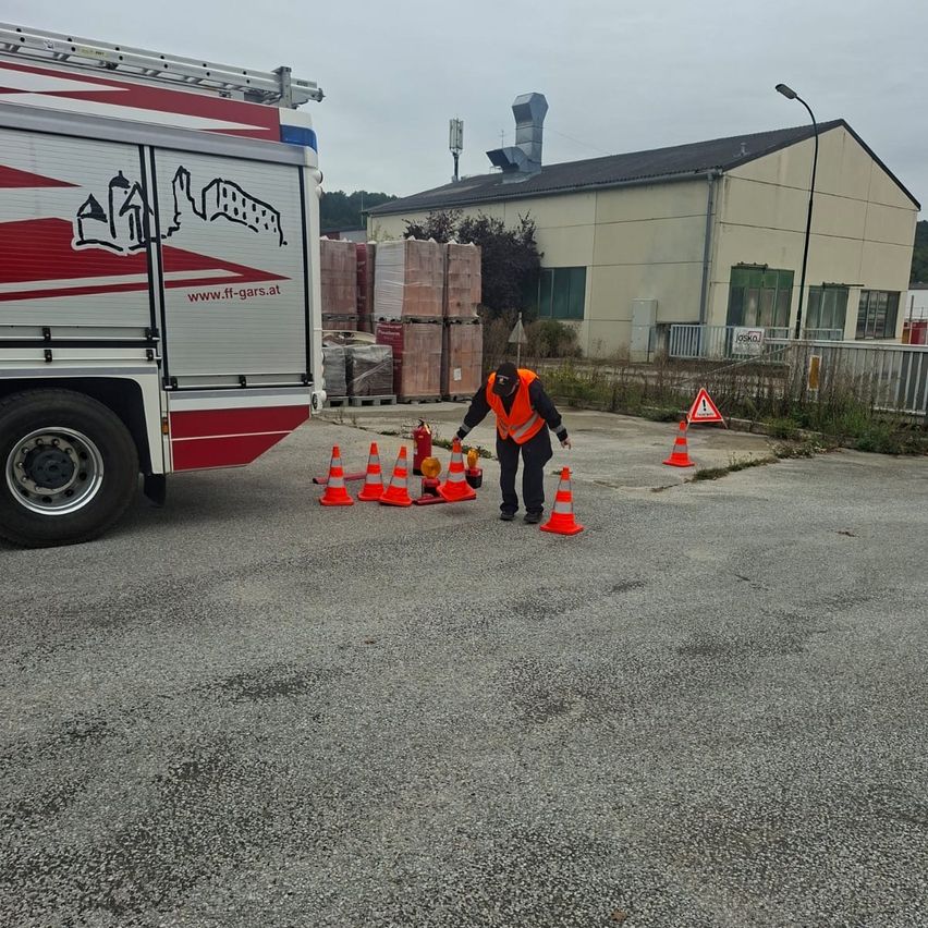 Eine Person in einer Warnweste und Hose steht vor einem Feuerwehrauto und Warnkegeln auf einem Parkplatz. Dahinter befindet sich ein Gebäude mit Lagerboxen.