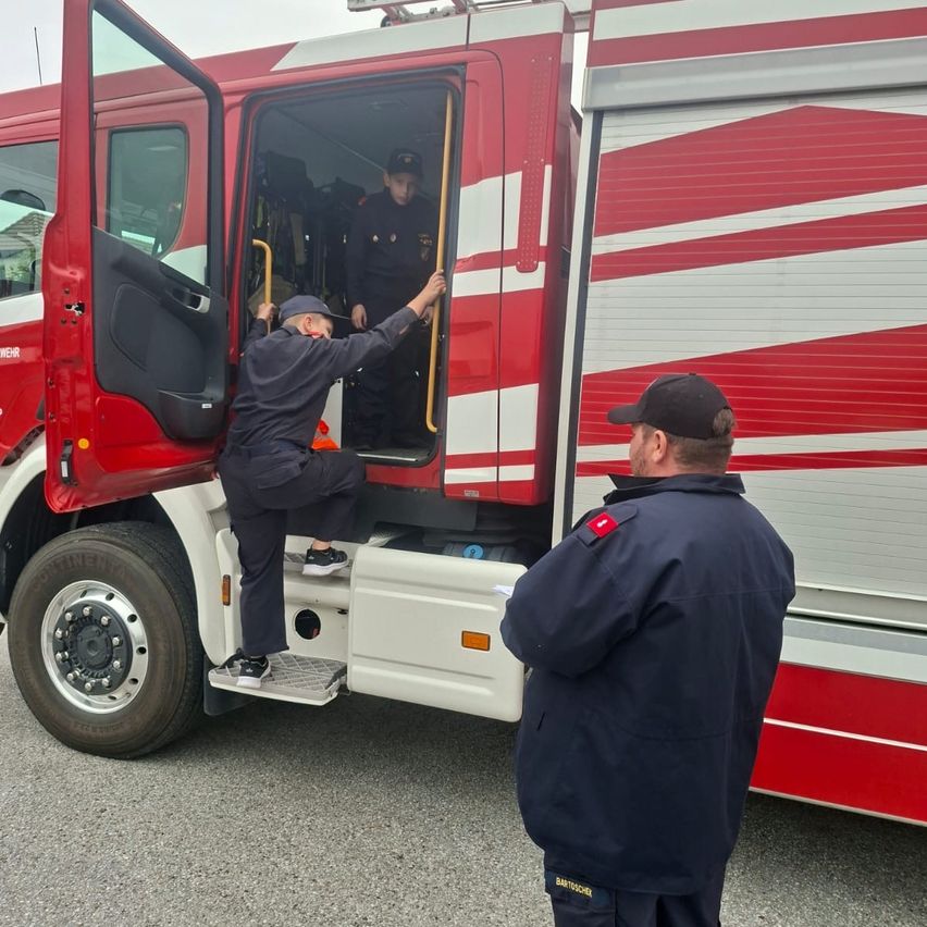 Zwei Feuerwehrleute in Uniform klettern in einen roten und weißen Feuerwehrwagen. Die Türen des Wagens sind offen. Ein Mann steht in der Nähe und beobachtet.