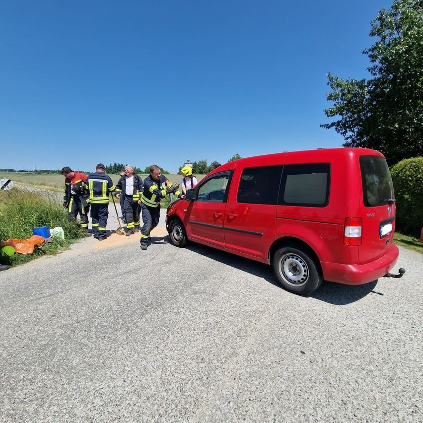 Ein roter Volkswagen Caddy ist am Straßenrand geparkt. Feuerwehrleute schieben das Fahrzeug. In der Nähe befinden sich einige orangefarbene und blaue Gegenstände.