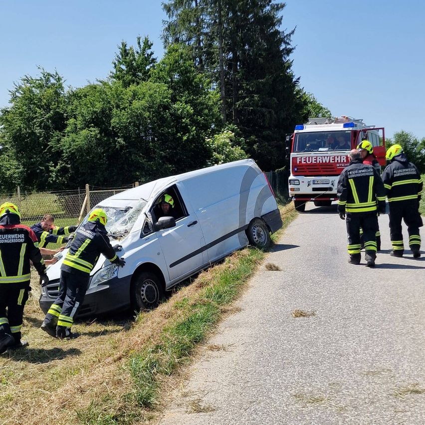 Ein weißer Van mit zerbrochener Windschutzscheibe steht am Straßenrand, umgeben von Feuerwehrleuten. Zwei Feuerwehrleute halten den Van fest.