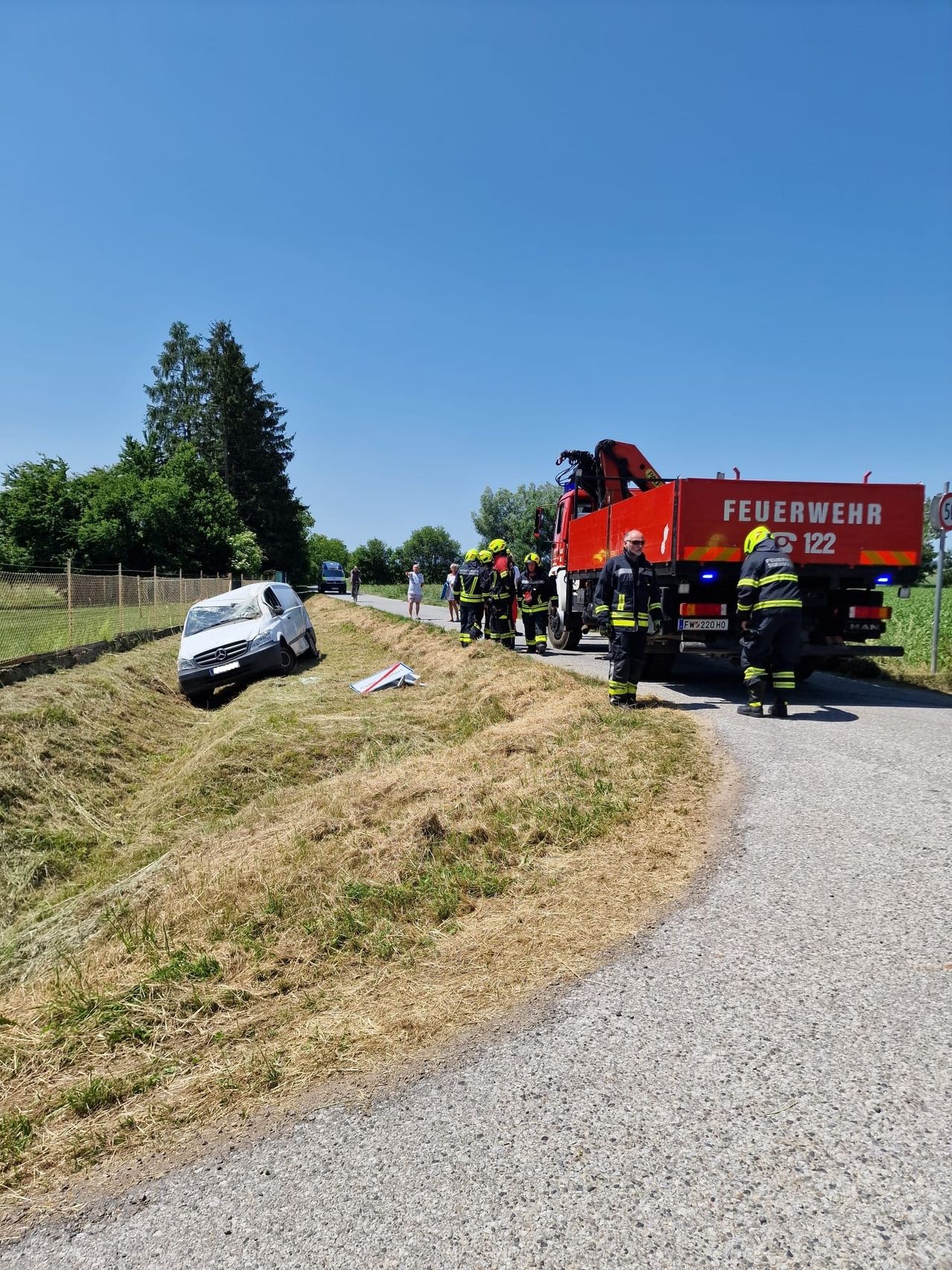 Feuerwehrleute in Schutzkleidung stehen neben einem roten Feuerwehrauto. Ein Auto ist an der Straßenseite beschädigt, mit einem Stück Trümmer in der Nähe.