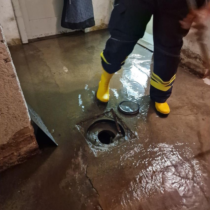 Eine Person in gelben Stiefeln steht über einem überfluteten Boden und untersucht ein wassergefülltes Loch.