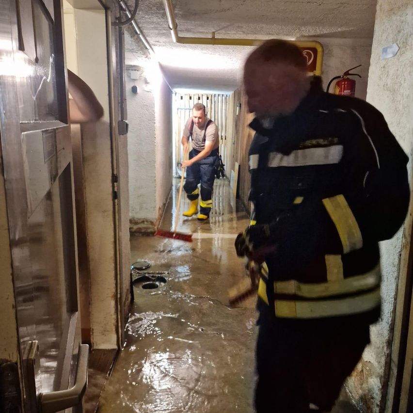 Zwei Männer in gelben Stiefeln und Latzhosen reinigen einen überfluteten Flur mit einem Besen. Einer schaut zurück, und ein Feuerlöscher ist an der Wand zu sehen.