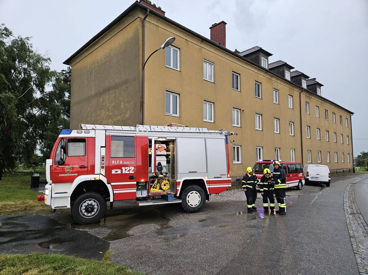 Ein Feuerwehrauto mit der Nummer 122 steht vor einem mehrstöckigen Gebäude mit mehreren Fenstern. Vier Feuerwehrleute stehen neben dem Auto.