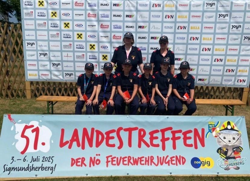 Eine Gruppe junger Feuerwehrleute posiert für ein Foto vor einem Banner mit der Aufschrift '51. LANDESTREFFEN DER NO FEVERWEHRJUGEND 3.-6. Juli 2025'. Dahinter zeigt ein Holzzaun verschiedene Logos.