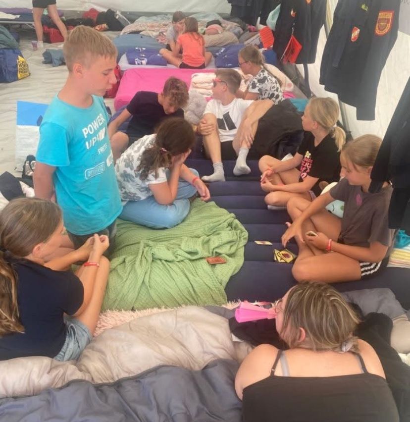In einem Zelt sitzen oder liegen mehrere Personen auf Matratzen, möglicherweise während eines Camps. Einige schauen nach unten, während andere in Gespräche vertieft sind.