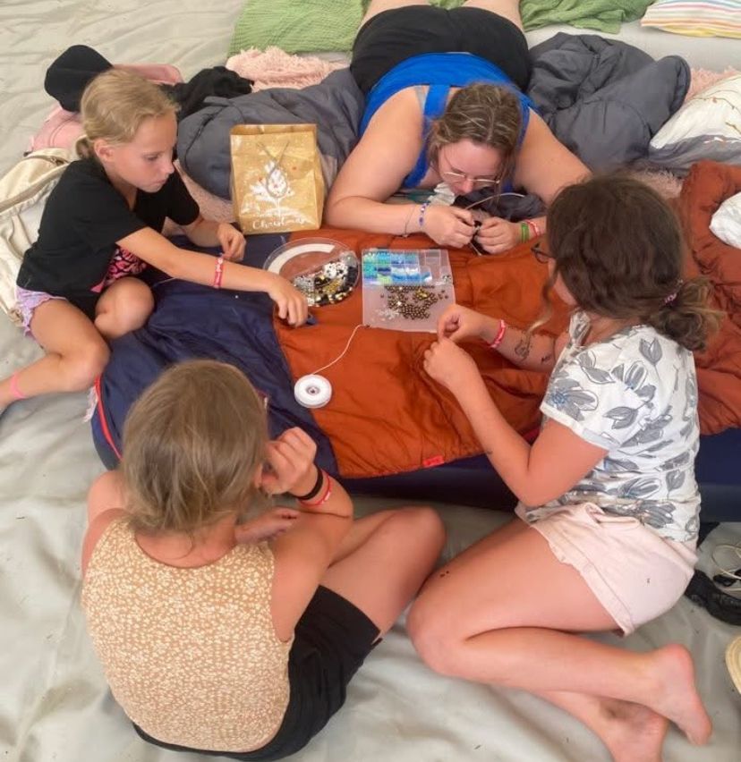 Eine Frau und drei Kinder sitzen auf einem Bett und arbeiten an einem Bastelprojekt. Die Frau liegt auf dem Bauch, und die Kinder sitzen kreuzbeinig. Sie haben Perlen, einen Behälter und eine Spule mit Faden.