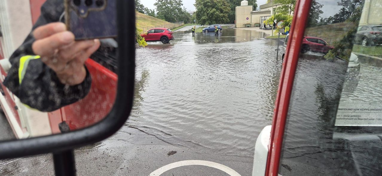 Eine überschwemmte Straße mit zwei Autos und zwei Personen im Wasser. Ein Gebäude befindet sich rechts.