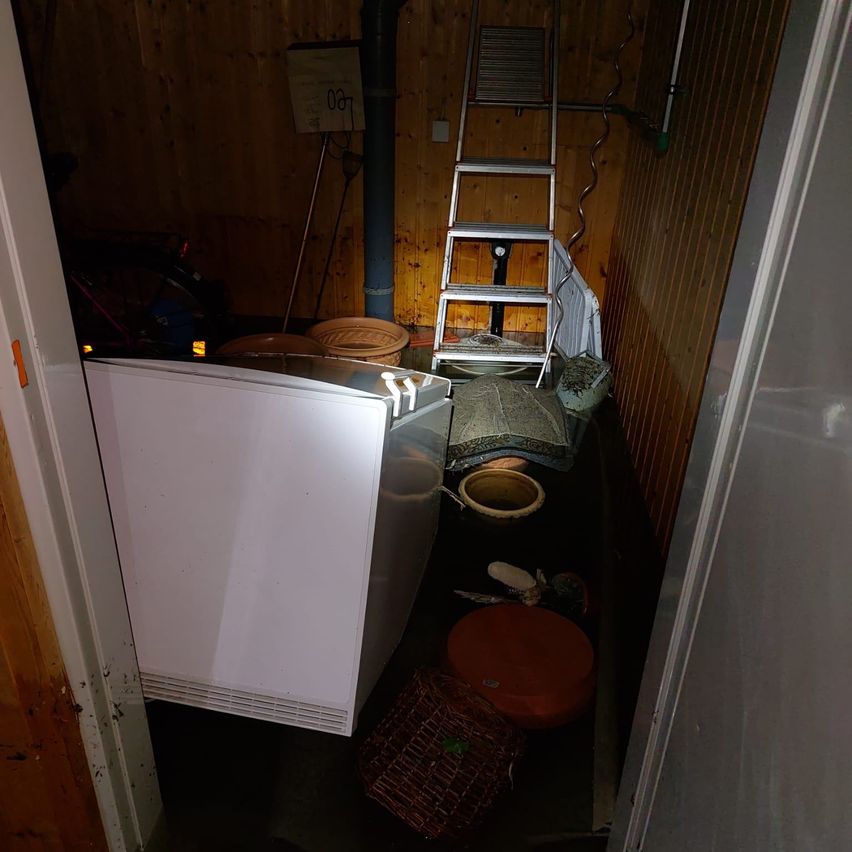 Ein überfüllter Lagerraum enthält einen Kühlschrank, eine Leiter und verschiedene Gegenstände wie Körbe und Schüsseln. Die Wände sind aus Holz.