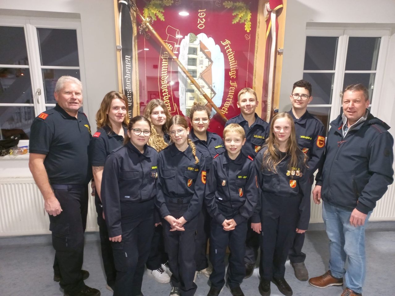 Eine Gruppe junger Menschen in Uniformen, wahrscheinlich Teil einer Jugendorganisation, posiert für ein Foto vor einem Banner mit deutschem Text. Sie werden von einigen Erwachsenen begleitet.