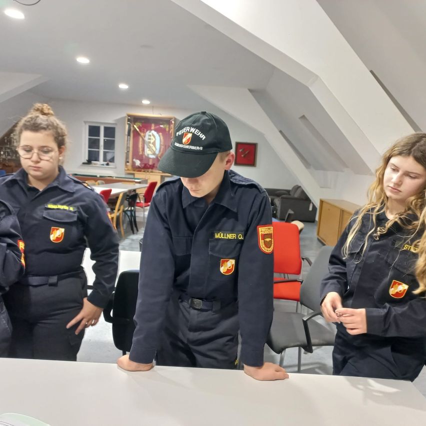 Drei Personen in Uniform sind in einem Raum mit einem Tisch. Der Mann in der Mitte beugt sich über den Tisch und die beiden Frauen stehen hinter ihm. Der Raum hat Stühle, einen Schreibtisch und einen Bilderrahmen an der Wand.