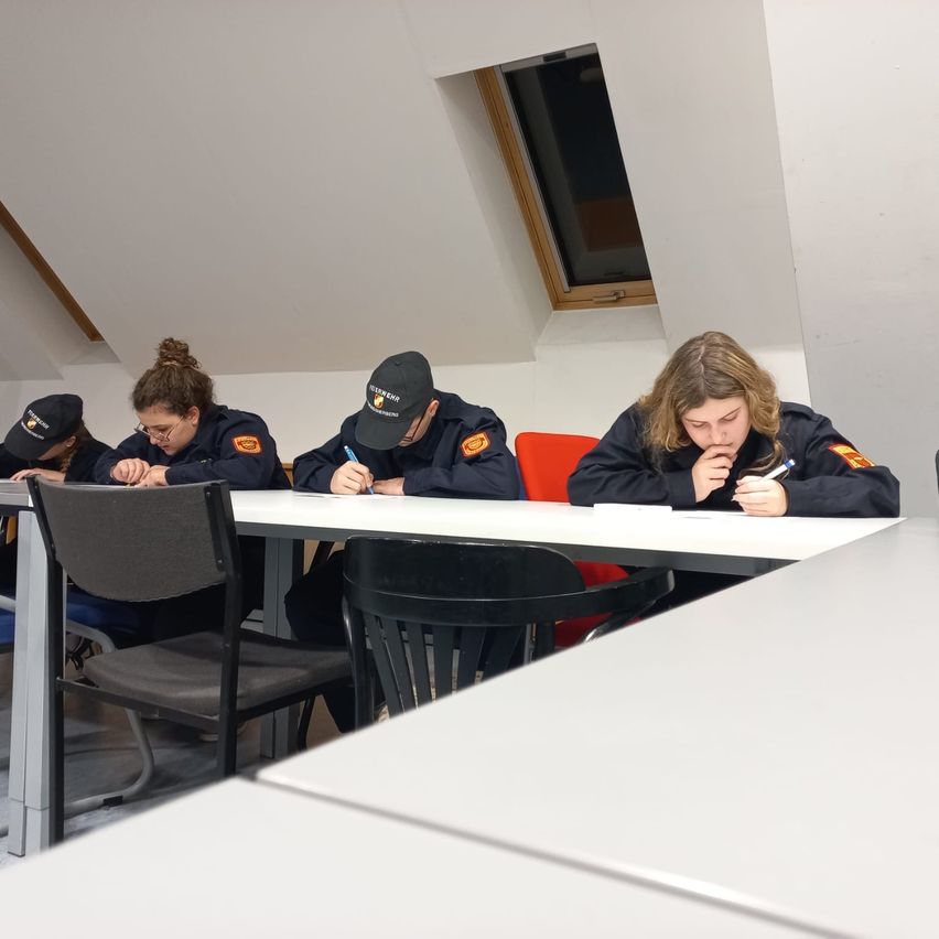 Vier Personen in Uniformen sitzen an einem Tisch und schreiben. Zwei sind Frauen, eine mit Brille, und zwei sind Männer, einer trägt einen Hut. Die Umgebung ist in Innenräumen mit einem Fenster im Hintergrund.