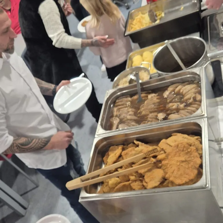 Eine Gruppe von Menschen steht um einen Buffettisch mit verschiedenen Speisen. Ein Mann in einem weißen Hemd hält einen Teller und Chopsticks. Das Buffet umfasst Würstchen, paniertes Essen und andere Gerichte.