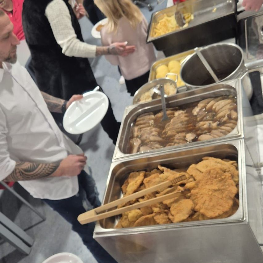 Eine Gruppe von Menschen steht um einen Buffettisch mit verschiedenen Speisen. Ein Mann in einem weißen Hemd hält einen Teller und Chopsticks. Das Buffet umfasst Würstchen, paniertes Essen und andere Gerichte.
