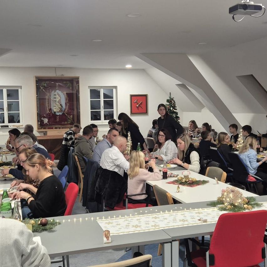 Eine Gruppe von Menschen isst an einem langen Tisch, der für Weihnachten geschmückt ist. Eine Frau steht und serviert Essen. Der Raum hat einen Projektor, einen Weihnachtsbaum und gerahmte Bilder an der Wand.