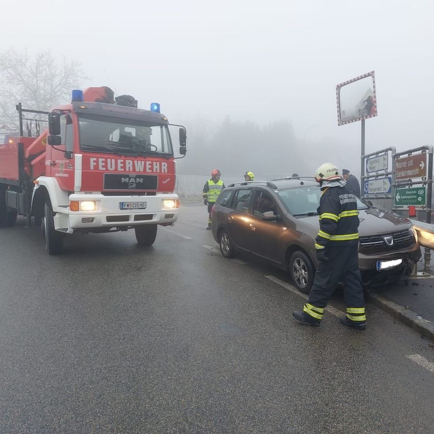 Ein Feuerwehrfahrzeug steht am Straßenrand in der Nähe eines beschädigten Autos. Ein Feuerwehrmann nähert sich dem Auto, während zwei andere in der Nähe stehen. Ein nebliger Hintergrund deutet auf schlechte Sicht hin.
