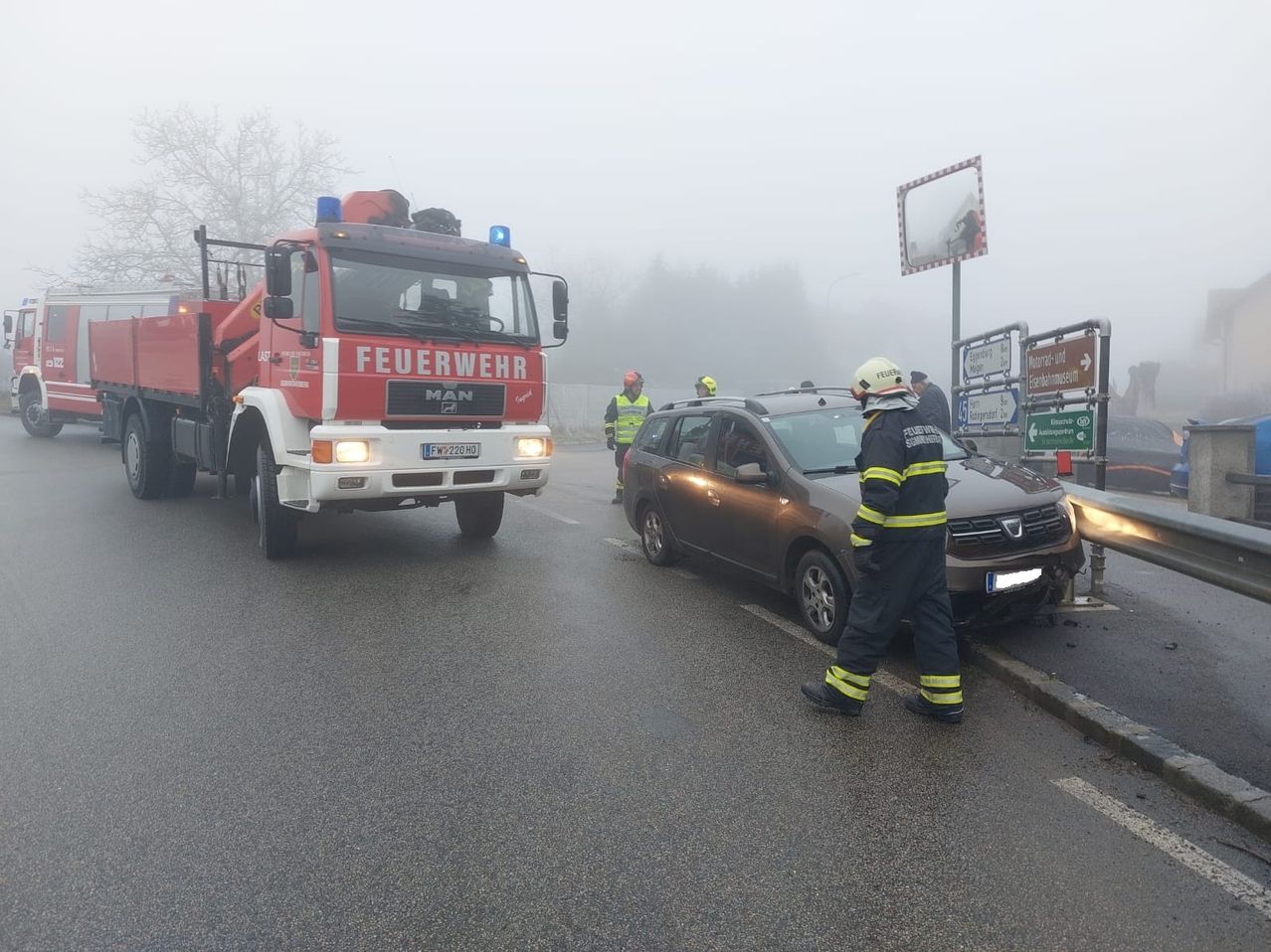 Ein Feuerwehrfahrzeug steht am Straßenrand in der Nähe eines beschädigten Autos. Ein Feuerwehrmann nähert sich dem Auto, während zwei andere in der Nähe stehen. Ein nebliger Hintergrund deutet auf schlechte Sicht hin.