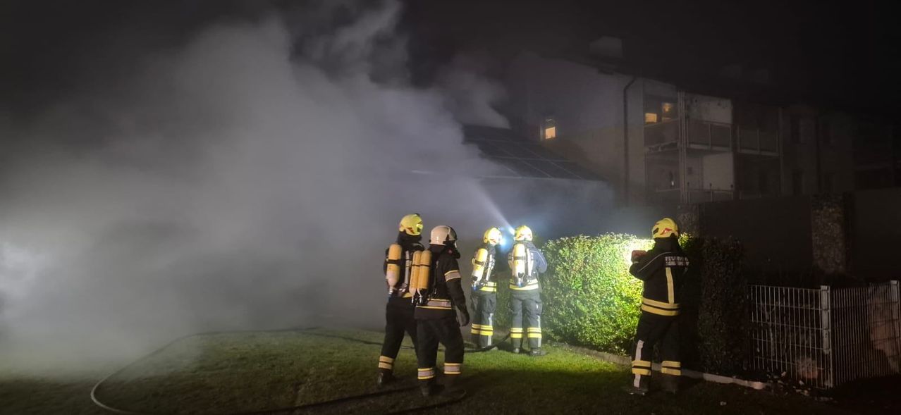 Feuerwehrleute in Schutzkleidung stehen auf einem Rasen und blicken auf ein Gebäude, aus dem Rauch aufsteigt. Sie sind von Taschenlampen beleuchtet.