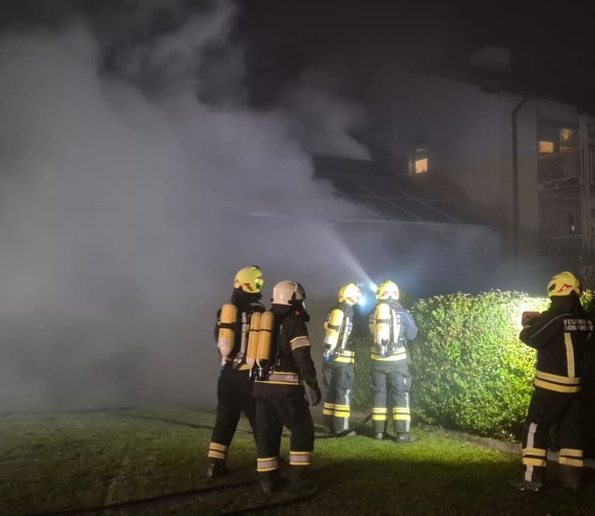 Feuerwehrleute in Schutzkleidung stehen auf einem Rasen und blicken auf ein Gebäude, aus dem Rauch aufsteigt. Sie sind von Taschenlampen beleuchtet.