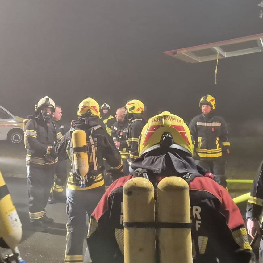 Eine Gruppe von Feuerwehrleuten in gelben und roten Uniformen steht bei Nacht auf einer Straße, wahrscheinlich in einer Besprechung, mit sichtbaren Feuerlöschern und Ausrüstung.