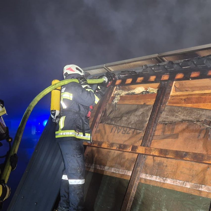 Ein Feuerwehrmann benutzt einen Schlauch, um ein Feuer auf dem Dach eines Gebäudes zu löschen. Der Feuerwehrmann ist in voller Schutzausrüstung gekleidet, einschließlich Helm, Jacke und Hose. Die Gebäudestruktur ist beschädigt, mit sichtbaren Brandflecken.