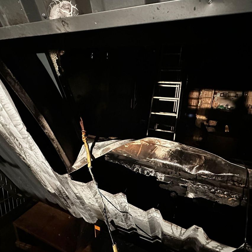 Eine Innenaufnahme mit einer beschädigten und freiliegenden Struktur. Ein weißer Tarp hängt über einem beschädigten Metallobjekt. Eine Leiter ist in der Nähe positioniert, und eine Person ist im Hintergrund sichtbar.