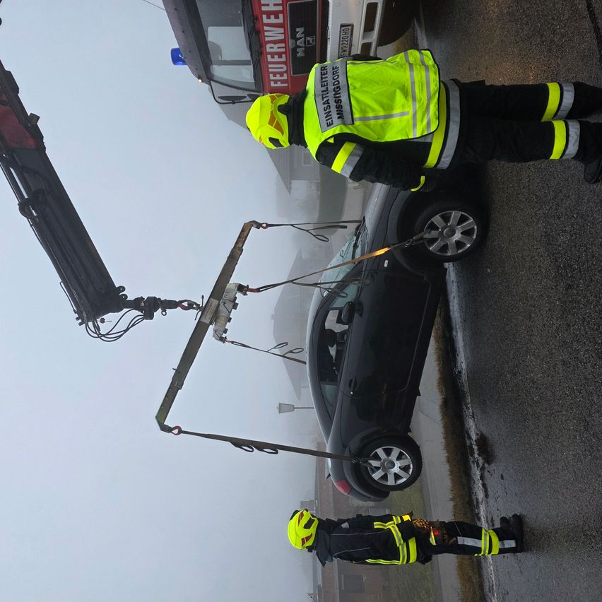 Ein Feuerwehrmann benutzt einen Kran, um ein Auto von der Straße zu entfernen. Ein anderer Feuerwehrmann hält das Seil. Das Auto wird vom Kran angehoben. Die Straße ist nass.