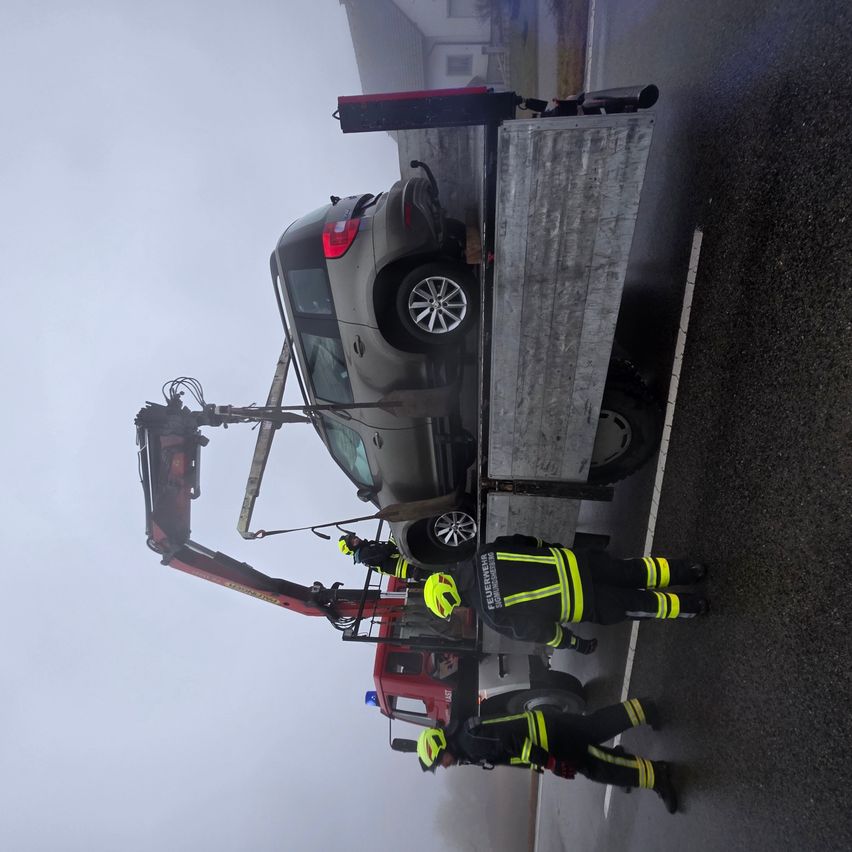 Rettungskräfte heben ein beschädigtes Fahrzeug auf einen Tieflader. Zwei Personen in Sicherheitsausrüstung helfen bei der Aktion.
