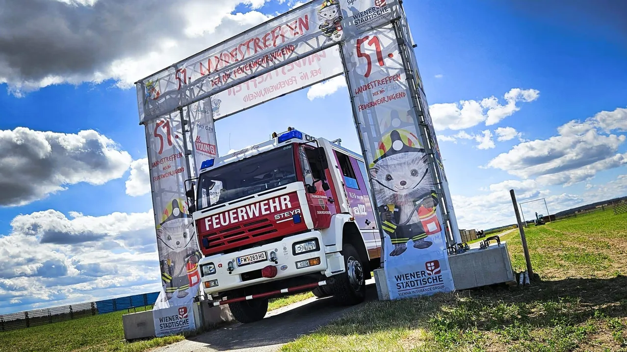 Ein Feuerwehrauto steht unter einem großen Torbogen mit Bannern, auf denen '57. Landstrassen' und 'Wiener Stadtische' steht. Das Auto hat ein Nummernschild, das 'FW 263 HO' liest. Der Himmel ist blau mit einigen Wolken.