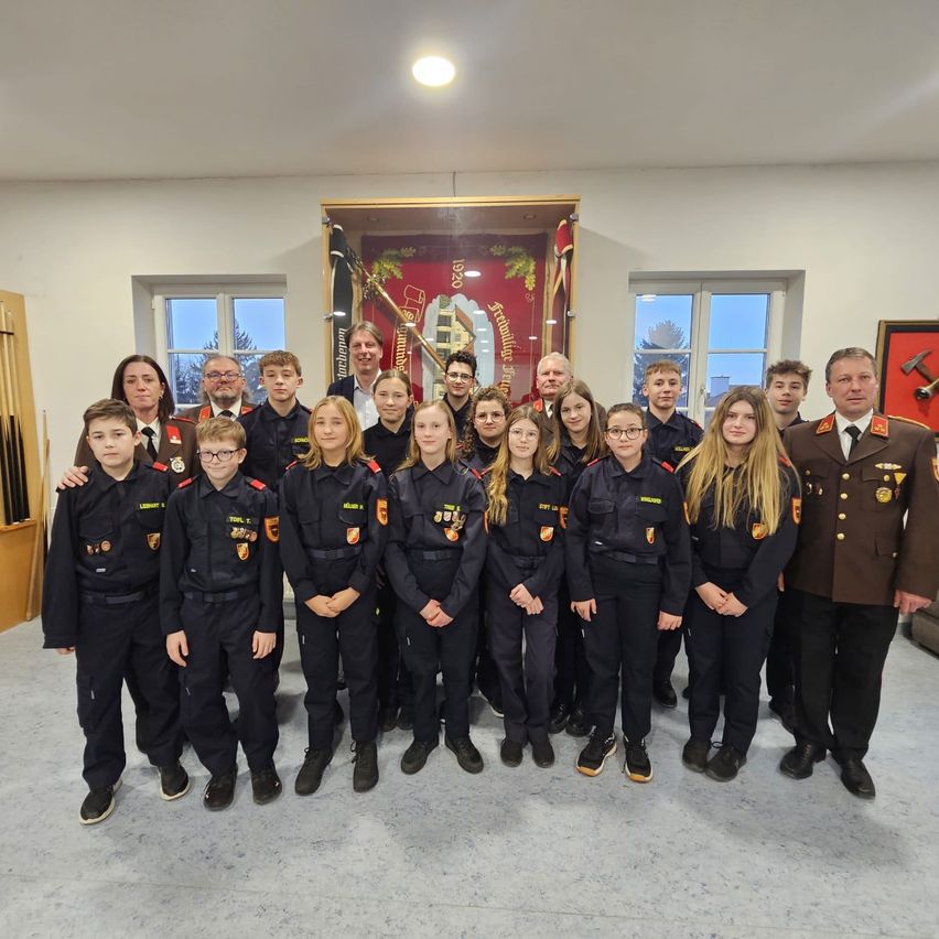 Eine Gruppe junger Menschen in Uniform, möglicherweise Kadetten, steht vor einem Banner und einem gerahmten Bild. Neben ihnen stehen zwei Erwachsene, ein Mann und eine Frau.