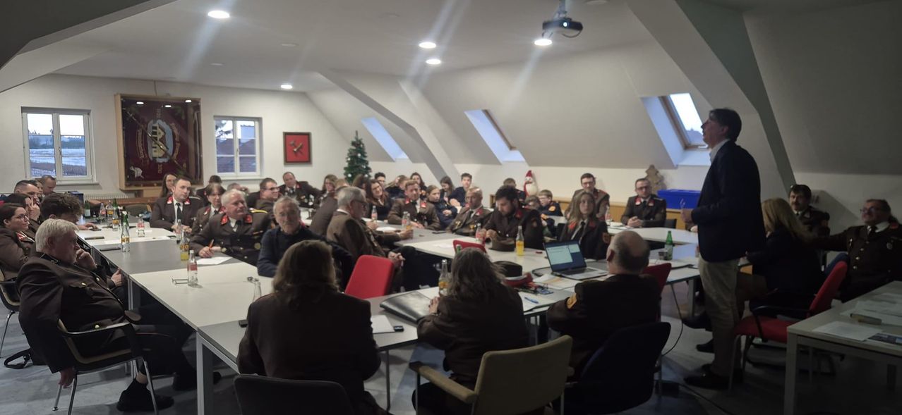 Eine Gruppe von Menschen in Uniform sitzt um einen langen Tisch in einem Besprechungsraum. Ein Weihnachtsbaum und ein gerahmtes Bild hängen an der Wand. Ein Projektor und Lampen sind an der Decke montiert.