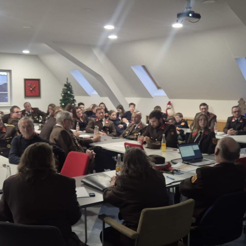 Eine Gruppe von Menschen in Uniform sitzt um einen langen Tisch in einem Besprechungsraum. Ein Weihnachtsbaum und ein gerahmtes Bild hängen an der Wand. Ein Projektor und Lampen sind an der Decke montiert.