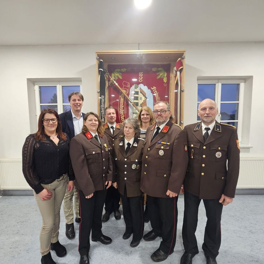 Eine Gruppe von Personen, sowohl Männer als auch Frauen, steht in einem Raum. Sie tragen Uniformen und scheinen für ein Foto zu posieren. Hinter ihnen befindet sich eine Flagge und ein Banner.