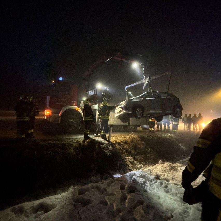 Feuerwehrleute verwenden einen Kran, um ein Auto aus dem Schnee bei Nacht zu heben. Eine Gruppe von Menschen steht herum.