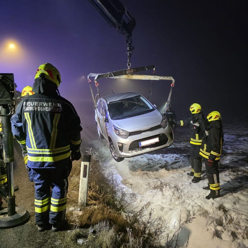 Drei Feuerwehrleute in gelben und schwarzen Uniformen heben einen silbernen Wagen mit einem Kran an. Das Auto steht am Rande einer verschneiten Straße, mit einem Pfosten in der Nähe.