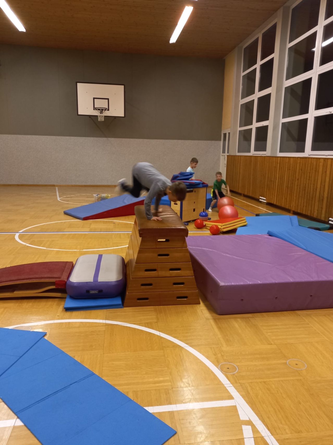 Drei Kinder spielen auf einem Basketballfeld und verwenden verschiedene farbige Matten und Bälle. Ein Kind führt einen Handstand auf einer Holzplattform durch.