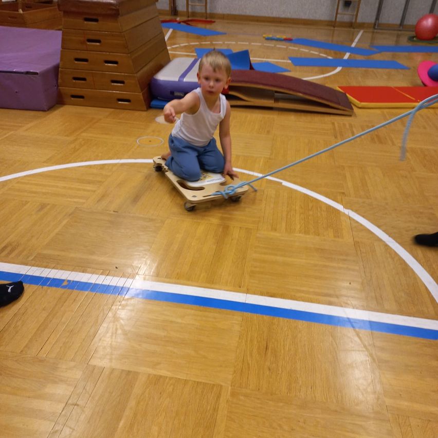 Ein junger Junge sitzt auf einem Skateboard im Innenbereich und zieht an einem Seil. Verschiedene Sportgeräte umgeben ihn auf einem Holzboden.
