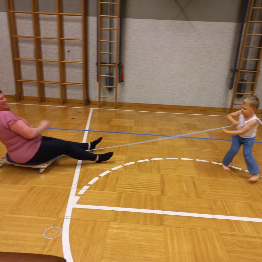 Eine erwachsene Frau und ein junger Junge spielen ein Spiel mit einem Seil auf einem Holzboden. Die Frau sitzt auf einem Schlitten, während der Junge auf der anderen Seite des Seils steht.