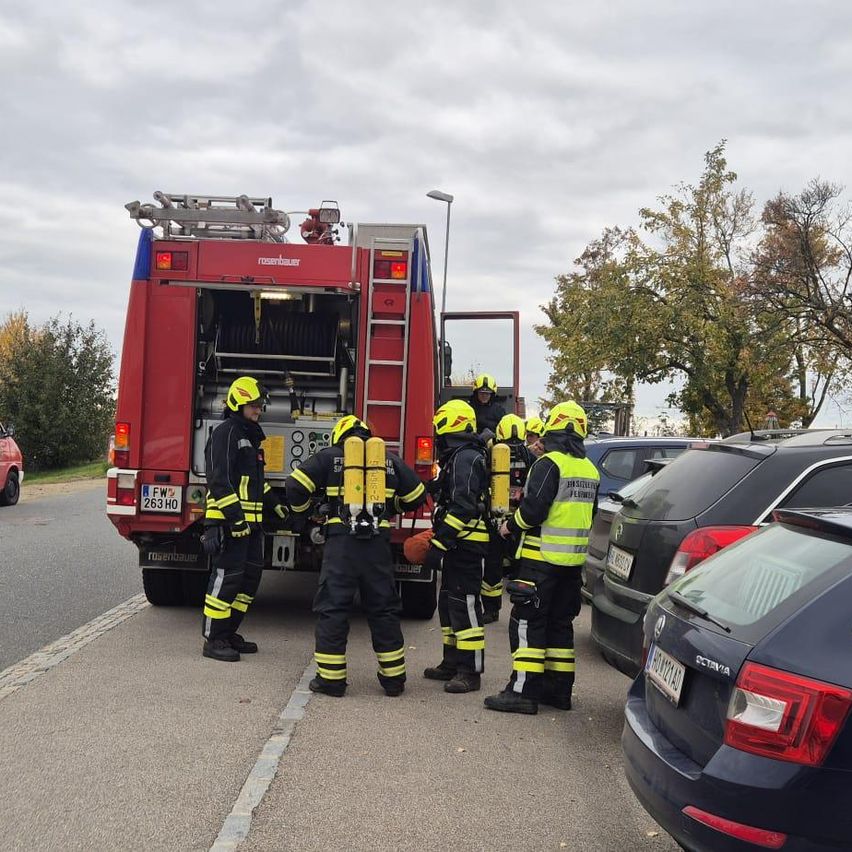 Fünf Feuerwehrleute stehen in der Nähe eines roten Feuerwehrwagens mit offener Tür auf einer Straße. Sie bereiten sich mit Helmen und Sauerstoffflaschen auf einen Notfall vor.
