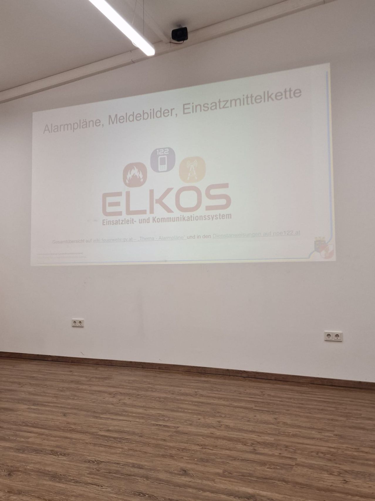 Eine Präsentationsfolie mit weißem Hintergrund zeigt ein Diagramm mit dem Titel 'ELKOS'. Das Diagramm enthält Symbole für Alarmpläne, Meldegeräte und Notfallkommunikationssysteme. Unter dem Diagramm steht der Text 'Einsatzleit- und Kommunikationssystem'. Der Raum hat weiße Wände und einen Holzboden.