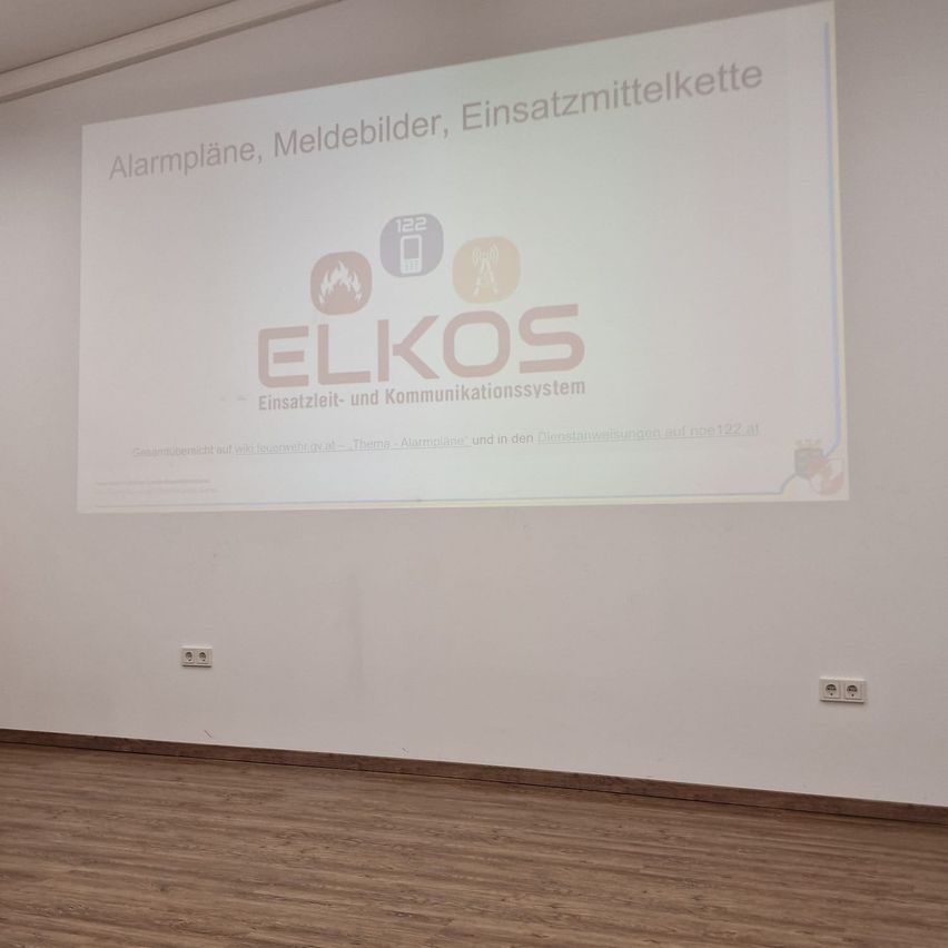 Eine Präsentationsfolie mit weißem Hintergrund zeigt ein Diagramm mit dem Titel 'ELKOS'. Das Diagramm enthält Symbole für Alarmpläne, Meldegeräte und Notfallkommunikationssysteme. Unter dem Diagramm steht der Text 'Einsatzleit- und Kommunikationssystem'. Der Raum hat weiße Wände und einen Holzboden.