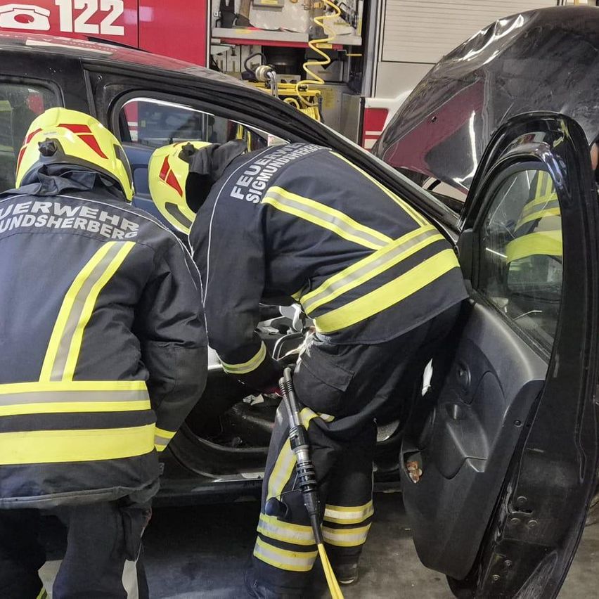Zwei Feuerwehrleute arbeiten an einem Auto mit offener Tür in einer Garage. Einer trägt einen gelben Helm und Handschuhe, der andere einen gelben und schwarzen Helm.