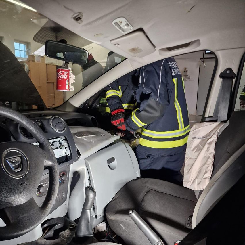 Ein Feuerwehrmann ist in einem Auto und repariert etwas am Armaturenbrett. Das Lenkrad befindet sich auf der linken Seite. Ein Feuerlöscher ist an der Wand dahinter montiert.