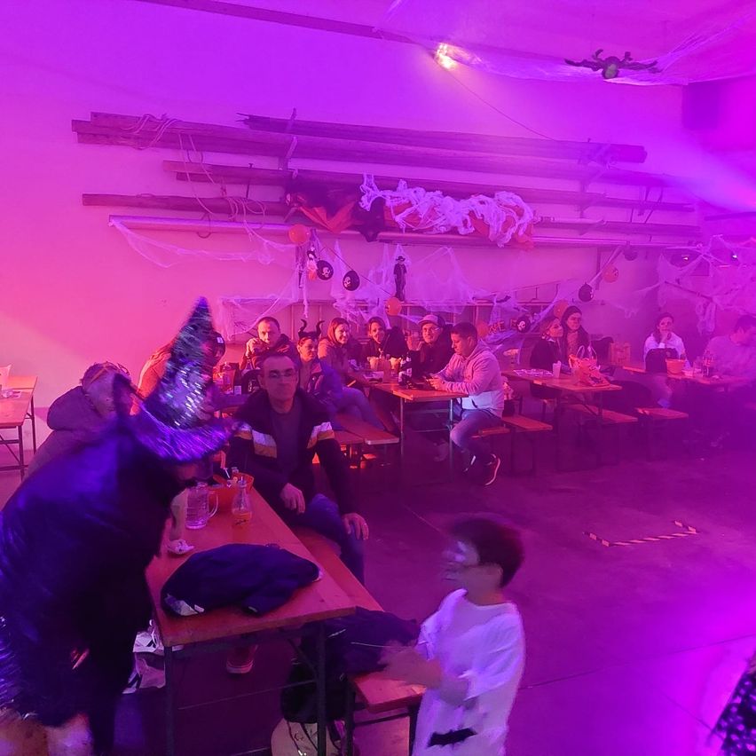 Eine Halloween-Party mit Menschen in Kostümen, die um Tische herum unter lila Lichtern sitzen. Ein Kind steht vor einem Tisch und schaut jemanden an.